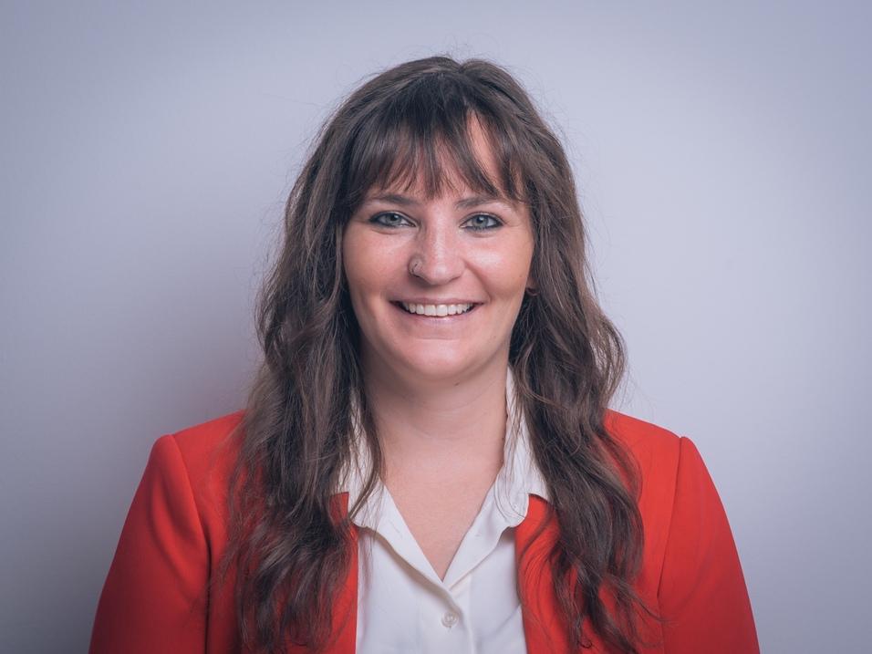 Porträt einer lächelnden Frau mit langen, lockigen Haaren, in einem roten Blazer und weißem Hemd, vor einfarbigem Hintergrund.