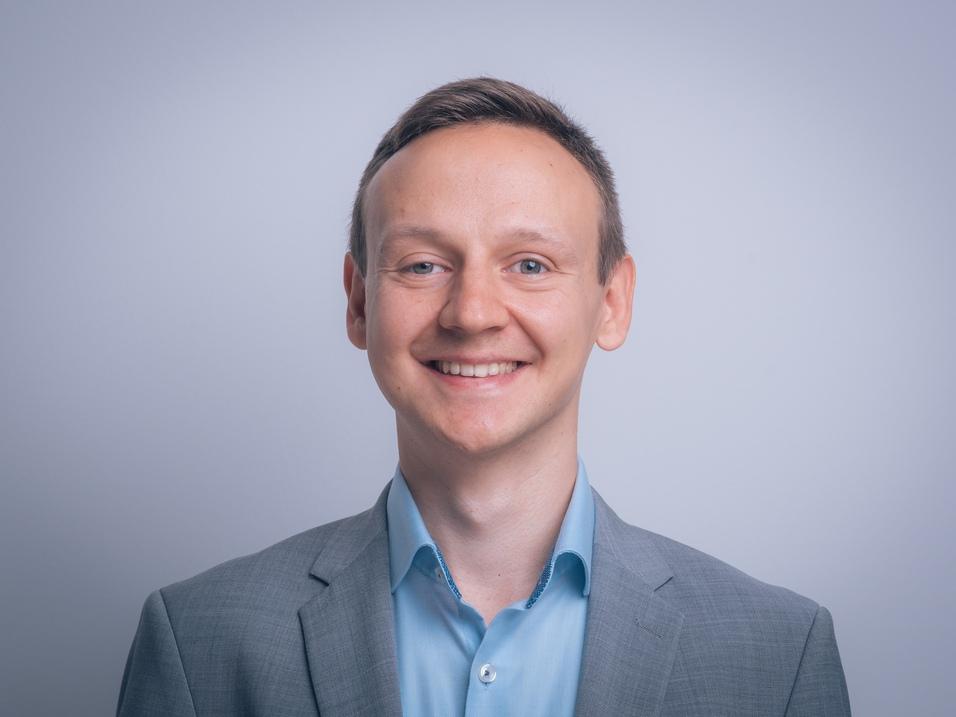 Porträt eines lächelnden Mannes in grauem Anzug und hellblauem Hemd vor einem hellen Hintergrund.